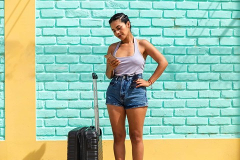 A young Latin woman stands against a colorful wall checking her phone next to luggage. The vibrant