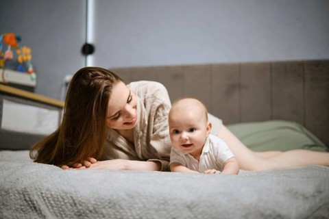 A mother is lying on a soft bed smiling at her baby who is crawling nearby. The room has a simple