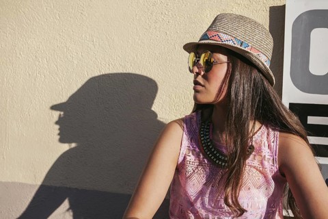 A stylish woman in a hat and sunglasses leans against a wall, her shadow forming a striking