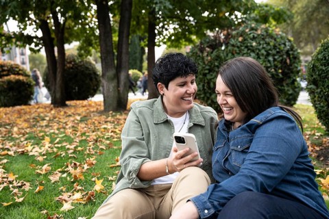 A joyful lesbian couple laughs together while looking at a phone, seated in a park with fallen