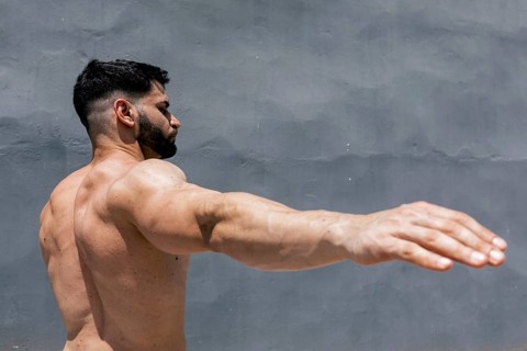 A fit, muscular man is seen stretching his arms against a gray wall outside. The image captures the