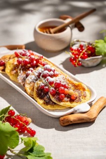 From above, a platter of golden pancakes topped with vibrant red currants sits on a light linen