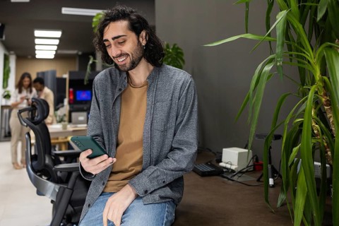 A professional at the office leans on a desk, smiling while engaging with a smartphone. In the