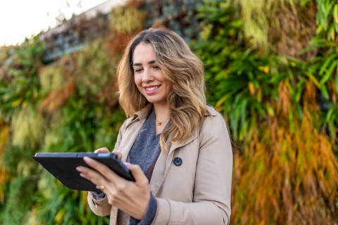 Woman smiling and wearing a trench coat while operating a digital tablet with her finger, standing