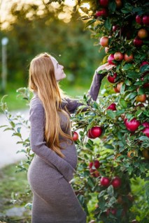 Beautiful pregnant woman with flowing red hair stands gracefully in an orchard, surrounded by lush