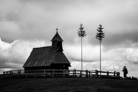 A black and white photograph of a traditional wooden chapel behind a fence on a ridge, with a lone