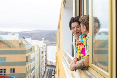 Lesbian couple lean out of a window on a vibrant city balcony, enjoying a peaceful moment together.