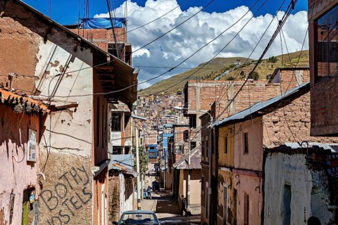 Narrow street with brick and plaster houses surrounded by mountains under blue sky with clouds, The