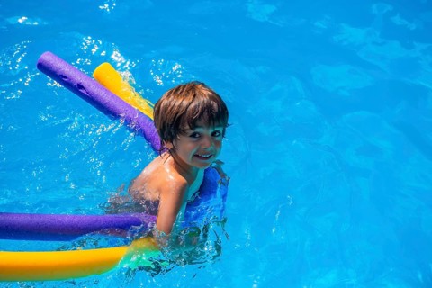 Little boy splashing joyfully in a bright blue pool with colorful foam noodles, smiling and playing
