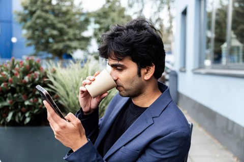 A indian man in a blue blazer sits outdoors, sipping coffee from a cup while scrolling through his