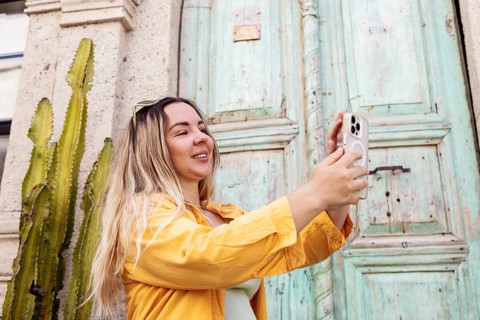A female tourist wearing a yellow shirt takes a selfie with a smartphone in front of an old,