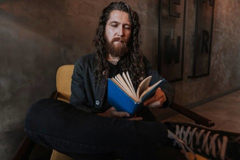 A man with long hair and a beard reads a blue book while sitting in a cozy chair of a cafe,