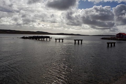 Abandoned and half-destroyed pier structures under a cloudy sky, near a small red hut on the shore,