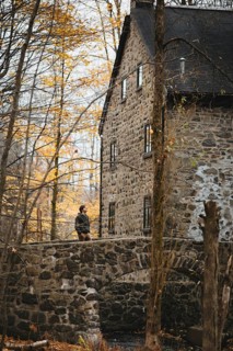A man walks on a stone bridge near an old stone building in an autumnal forest in Quebec, Canada.