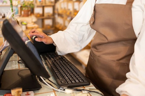 A store clerk in an organic supermarket adjusts the point of sale system, wearing a brown apron.