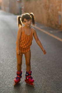 A young girl in a striped outfit enjoys rollerblading on a sunlit street in Santa Maria del Cami,
