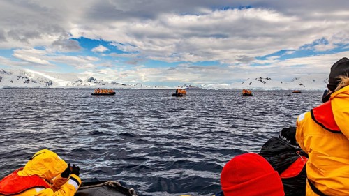 People in orange and yellow jackets on a boat navigating between icebergs under a cloudy sky,