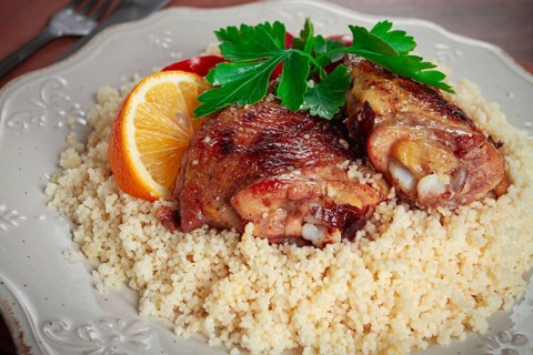 Couscous is served on a plate with pieces of chicken on top. Lemon slices and parsley decorate the