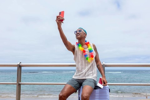 An LGBTQIA+ man sitting on a suitcase on the beach, adorned with a colorful flower necklace, takes