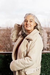 A smiling woman with white hair enjoys the view from her balcony, dressed warmly in a cozy jacket.