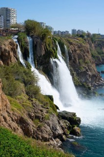 Dynamic waterfall flows into the sea surrounded by rocks and cityscape in the background, Lower