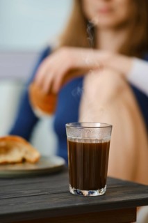 Coffee glass with unrecognizable blurred woman sitting in the kitchen with croissant in hand, focus