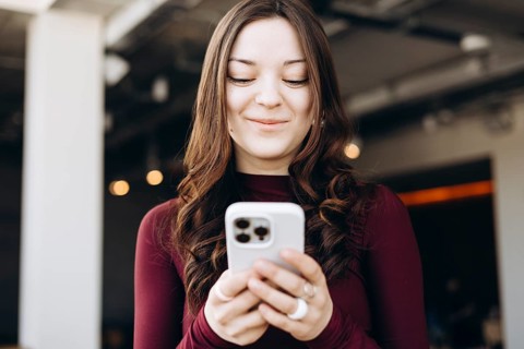 A young female student with long hair stands indoors, focused on her smartphone. She wears a