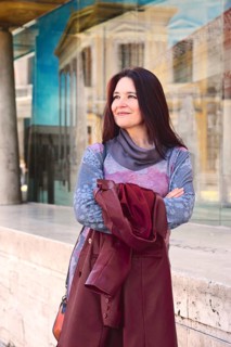 A woman stands confidently outdoors, wearing a colorful sweater and holding a maroon jacket. She is
