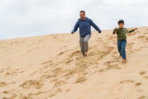 A joyful father and son running barefoot down the dune of Pilat on a cloudy day, sharing a moment