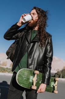 A stylish man in a leather jacket drinks coffee while holding a skateboard under the bright blue