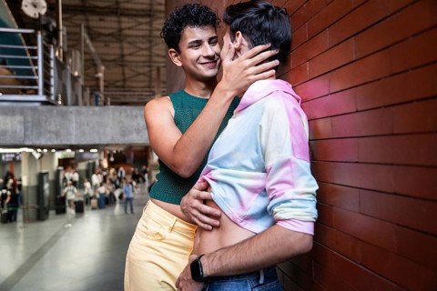 A gay couple shares a tender moment in a train station, showcasing love and diversity. The vibrant