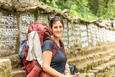 Smiling woman with a backpack trekking along a lush trail near Everest, surrounded by stone walls