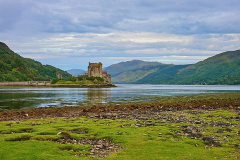 View of a historic riverside castle with mountainous backdrop and cloudy sky, Eilean Donan Castle,