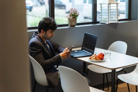 A indian man in a suit multitasks using a smartphone and laptop in a hotel cafeteria at a