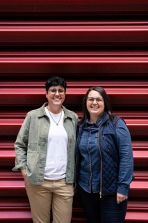 A joyful lesbian couple poses in casual attire against a red backdrop, showcasing their happiness