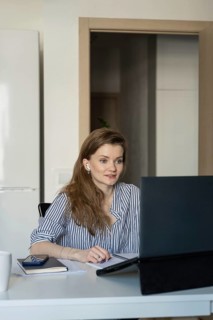 A woman in a striped shirt works on a laptop at home, engaging in a virtual meeting. She appears
