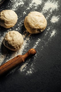 Three dough balls rest on a dark, floured surface, accompanied by a wooden rolling pin, ready for