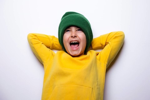 A cheerful child dressed in a vibrant yellow hoodie and a green beanie, arms resting behind head,