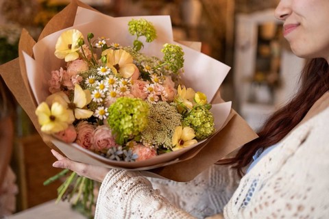 A woman holding a beautifully arranged bouquet of flowers, featuring daisies, roses, and greens, in