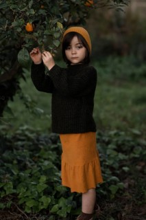 A young child in a mustard hat and skirt stands by an orange tree, exploring nature. They are