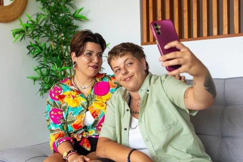 A cheerful lesbian couple sitting on a sofa, taking a selfie with a smartphone. The cozy living