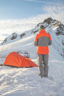 A person stands near a bright orange tent amidst snowy mountains under a clear sky. The scene
