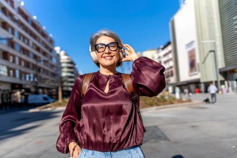 Senior woman strolling through a sunny city street, smiling and listening to music with headphones,