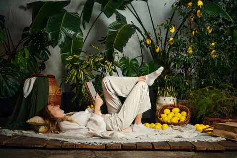 A woman is lying on the floor and reading a book. She is surrounded by plants and baskets of lemons