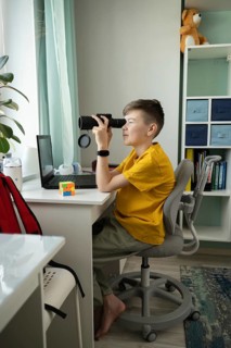 A curious boy in a yellow shirt sits barefoot at his desk using a monocular. Surrounded by a