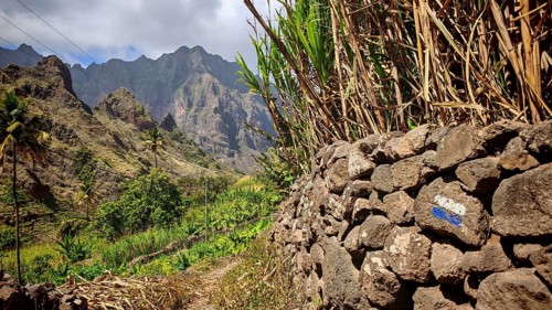 Stone wall with hiking trail, blue and white marker, behind it majestic mountains under cloudy sky,