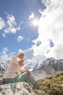 A woman sits on a rocky ledge, smiling warmly, against a backdrop of snow-covered Everest peaks,