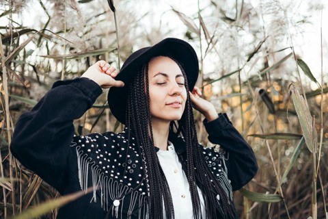 A woman with braids stands serenely in a field at sunset, wearing a stylish hat and closing her