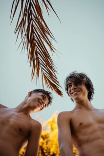 Two teen boys enjoy a sunny day outdoors, laughing under palm tree leaves against a clear sky.