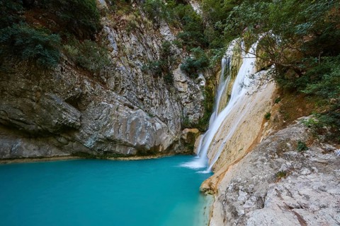 A waterfall flows over rocks into a tranquil turquoise pool surrounded by thick vegetation,